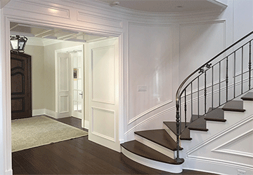 Angled view of the foot of a curved hardwood staircase and glimpse into a spacious hallway, with paneled white walls