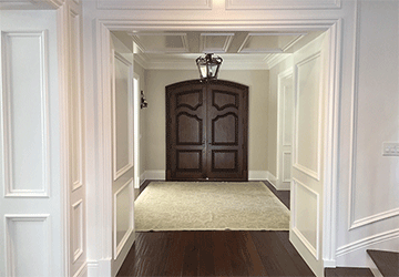 Square view through a large white-paneled hallway entry, looking to tall, curved, hardwood double doors on the far side of the next room